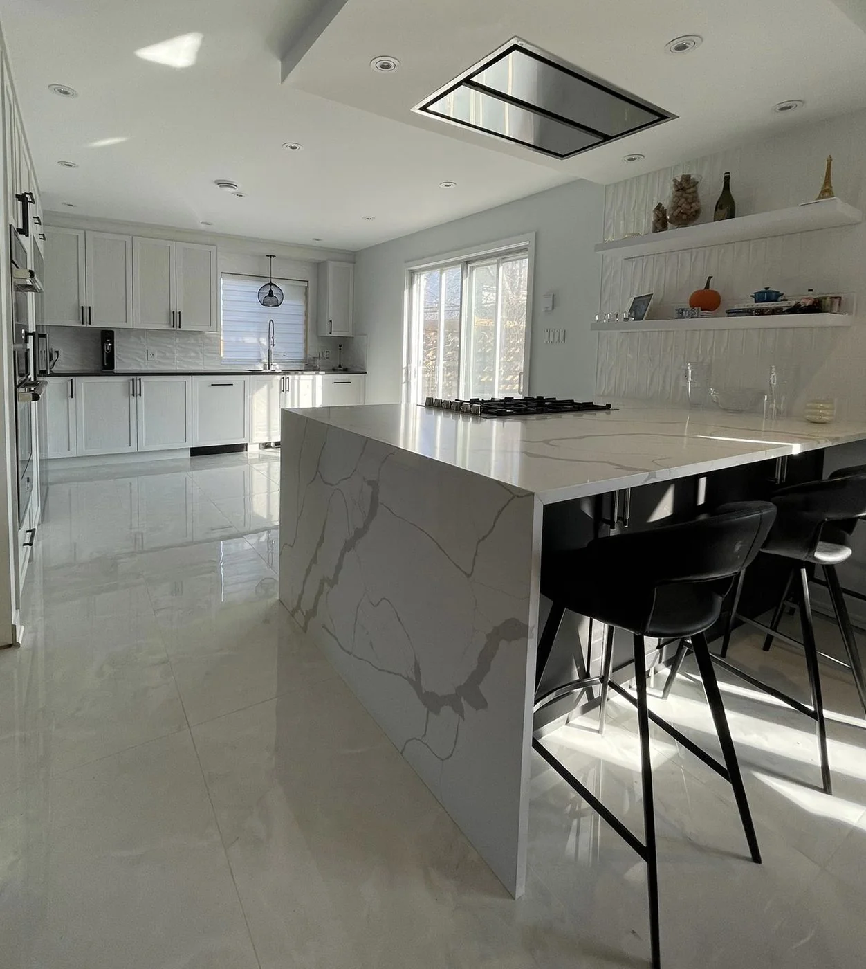 Bright white kitchen with a large waterfall quartz island in grey veined stone, black bar stools and a ceiling integrated hood