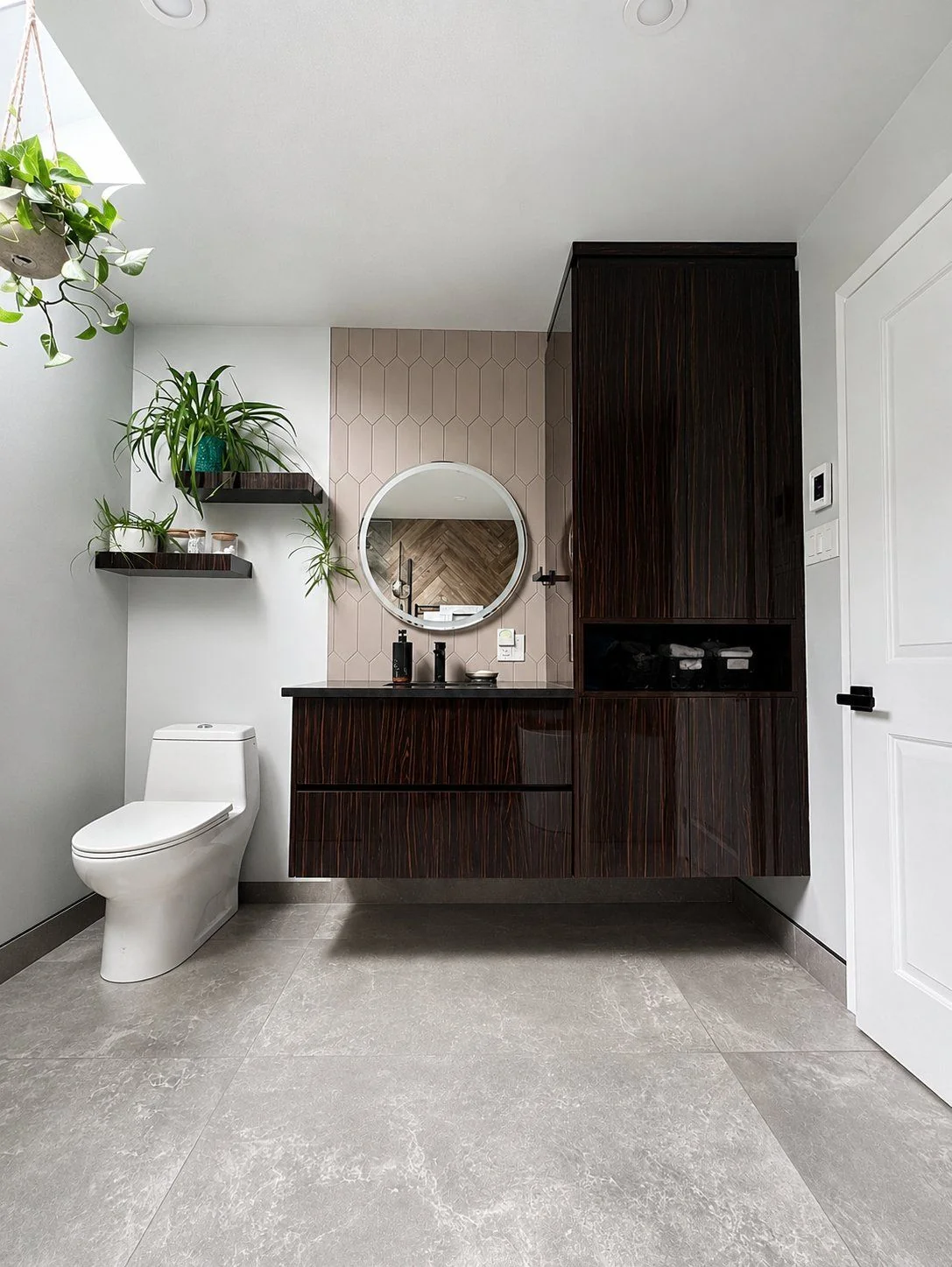 Vue large d’une salle de bain avec vanité murale en bois foncé, miroir rond et plancher de céramique grand format, par Limakdéco