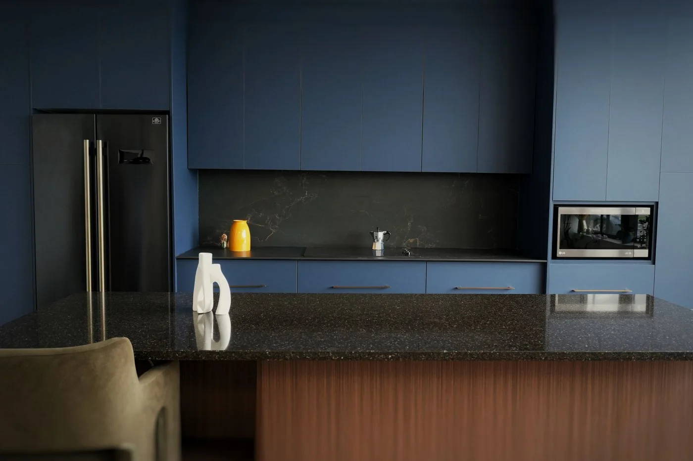 Portes d’armoires bleues avec comptoir de quartz foncé moucheté, four micro-ondes intégré et réfrigérateur en acier, par Limakdéco
