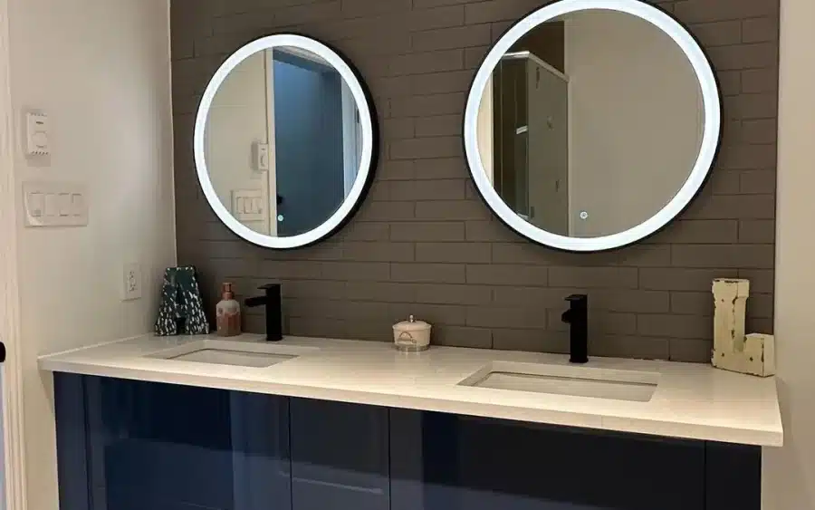 Wall mounted navy gloss double vanity with two illuminated round mirrors on a dark brick tiled wall, by Limakdeco