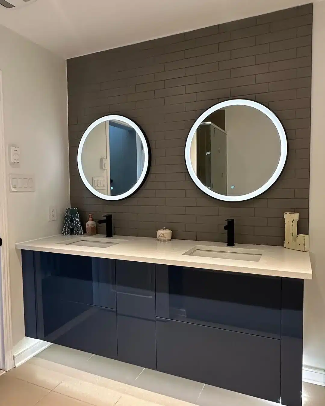 Wall mounted navy gloss double vanity with two illuminated round mirrors on a dark brick tiled wall, by Limakdeco